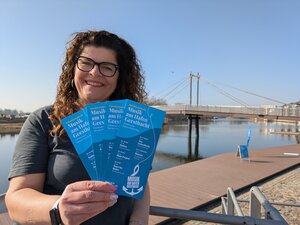 Eine l&auml;chelnde Frau mit braunen Locken und Brille steht an der Hafenpromenade in Geesthacht. Sie h&auml;lt f&auml;cherf&ouml;rmig mehrere blaue Flyer f&uuml;r die Veranstaltungsreihe &raquo;Musik am Hafen Geesthacht&laquo; in die Kamera. Im Hintergrund erstreckt sich die Elbe unter einem klaren blauen Himmel, &uuml;berspannt von der modernen Hafenbr&uuml;cke. Die Szene ist hell und sonnig.