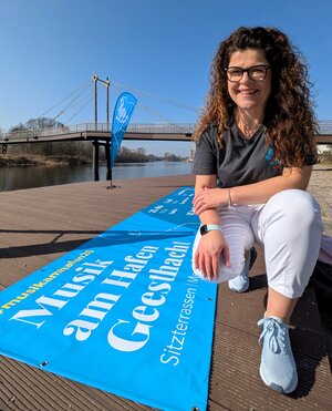 Bild vergr&ouml;&szlig;ern: Eine l&auml;chelnde Frau mit langen braunen Locken und Brille kniet auf einer h&ouml;lzernen Uferpromenade in Geesthacht. Sie tr&auml;gt ein graues T-Shirt und eine wei&szlig;e Hose. Vor ihr auf dem Boden liegt ein gro&szlig;es, leuchtend blaues Banner mit der Aufschrift &raquo;Musik am Hafen Geesthacht&laquo;. Im Hintergrund sind die Elbe, eine moderne Fu&szlig;g&auml;ngerbr&uuml;cke und eine blaue Beachflag unter strahlend blauem Himmel zu sehen.