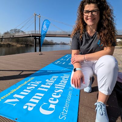 Eine l&auml;chelnde Frau mit langen braunen Locken und Brille kniet auf einer h&ouml;lzernen Uferpromenade in Geesthacht. Sie tr&auml;gt ein graues T-Shirt und eine wei&szlig;e Hose. Vor ihr auf dem Boden liegt ein gro&szlig;es, leuchtend blaues Banner mit der Aufschrift &raquo;Musik am Hafen Geesthacht&laquo;. Im Hintergrund sind die Elbe, eine moderne Fu&szlig;g&auml;ngerbr&uuml;cke und eine blaue Beachflag unter strahlend blauem Himmel zu sehen.