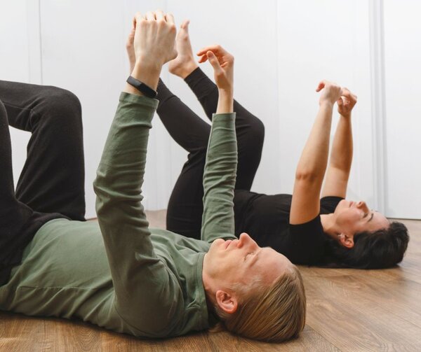 Zwei Personen, ein Mann im Vordergrund und eine Frau dahinter, liegen auf dem R&uuml;cken auf einem hellen Holzboden in einem hellen Raum. Sie f&uuml;hren achtsame Bewegungen im Rahmen einer Feldenkrais-Lektion aus.
Vordergrund: Ein Mann mit blondem Haar tr&auml;gt ein lang&auml;rmeliges, olivgr&uuml;nes Shirt und eine dunkle Hose. Seine Augen sind geschlossen, w&auml;hrend er seine Arme senkrecht nach oben streckt und die H&auml;nde entspannt h&auml;lt. Seine Beine sind angewinkelt.
Hintergrund: Eine Frau mit dunklen Haaren in schwarzer Sportkleidung f&uuml;hrt eine &auml;hnliche Bewegung aus. Ihre Arme und Beine sind ebenfalls vom Boden abgehoben und in die Luft gestreckt.