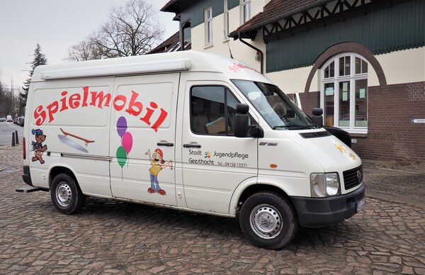 Bild vergr&ouml;&szlig;ern: Ein wei&szlig;er Kleintransporter der Stadtjugendpflege Geesthacht, beschriftet als &raquo;Spielmobil&laquo;. Die Seitenwand des Fahrzeugs ist mit dem gro&szlig;en roten Schriftzug &raquo;Spielmobil&laquo; sowie bunten Illustrationen eines B&auml;ren, eines Skateboards, Luftballons und einer Comicfigur verziert. Der Wagen parkt auf einer Kopfsteinpflasterstra&szlig;e vor einem historischen Geb&auml;ude mit wei&szlig; gerahmten Rundbogenfenstern. Auf der Fahrert&uuml;r befinden sich das Logo der Stadt Geesthacht und eine Telefonnummer.