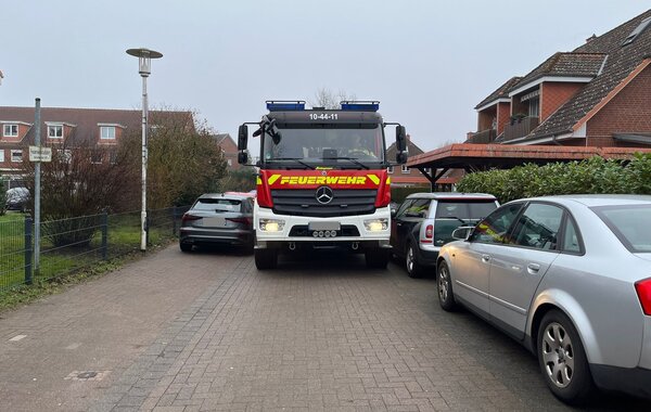 Frontalansicht eines gro&szlig;en roten Feuerwehrfahrzeugs, das auf einer gepflasterten Wohnstra&szlig;e steht. Mehrere parkende Autos, insbesondere ein silberner Pkw auf der rechten Seite, verengen die Fahrbahn erheblich. Das Feuerwehrfahrzeug mit eingeschaltetem Abblendlicht und der Kennung &raquo;10-44-11&laquo; scheint Schwierigkeiten zu haben, die enge Passage zu passieren. Im Hintergrund sind Wohngeb&auml;ude mit Backsteinfassaden zu sehen. Das Bild verdeutlicht die Problematik von durch parkende Autos blockierten Rettungswegen.