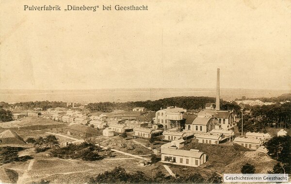 Bild vergr&ouml;&szlig;ern: Historische Postkarte in Sepia-Optik mit der Aufschrift &raquo;Pulverfabrik ,D&uuml;neberg' bei Geesthacht&laquo;. Die Luftaufnahme zeigt das weitl&auml;ufige Werksgel&auml;nde im Jahr 1915. Zu sehen sind zahlreiche flache Fabrikgeb&auml;ude, Schuppen und ein markanter, hoher Industrieschornstein, eingebettet in eine bewaldete H&uuml;gellandschaft. Im Hintergrund erstreckt sich eine weite, flache Ebene. Unten rechts ist ein modernes Wasserzeichen des &raquo;Geschichtsverein Geesthacht&laquo; eingef&uuml;gt.