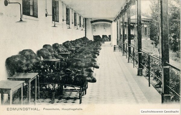 Bild vergr&ouml;&szlig;ern: Historische Schwarz-Wei&szlig;-Postkarte aus dem Jahr 1907 mit der Aufschrift &raquo;EDMUNDSTHAL. Frauenheim, Hauptliegehalle.&laquo; Die Aufnahme zeigt eine lange, &uuml;berdachte und zur Seite offene Veranda mit einer Reihe von gepolsterten Liegest&uuml;hlen und kleinen Beistelltischen f&uuml;r Freiluft-Liegekuren. An der R&uuml;ckwand h&auml;ngen elektrische Lampen. Rechts bietet die Halle einen Blick ins Gr&uuml;ne und auf einen h&ouml;lzernen Pavillon. Unten rechts befindet sich der Hinweis &raquo;Geschichtsverein Geesthacht&laquo;.