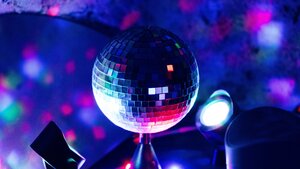 Eine Nahaufnahme einer funkelnden Discokugel, die von einem hellen Scheinwerfer angestrahlt wird. Sie steht im Zentrum einer dunklen Partyumgebung. Der Hintergrund ist erf&uuml;llt von einem bunten, verschwommenen Meer aus blauen, pinken, lilafarbenen und gr&uuml;nen Lichtpunkten. Die Atmosph&auml;re wirkt lebendig, fr&ouml;hlich und energiegeladen, als w&auml;re man mitten auf einer Tanzfl&auml;che. Ein weiterer kleinerer Scheinwerfer ist rechts im Bild zu sehen.