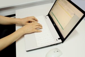 Eine Person tippt auf der Tastatur eines wei&szlig;en LG-Laptops, der auf einem hellen Schreibtisch steht. Auf dem Bildschirm ist ein Tabellenkalkulationsprogramm ge&ouml;ffnet, und im Vordergrund liegt eine wei&szlig;e Computermaus.