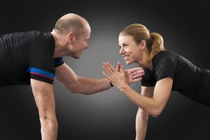 Ein Mann und eine Frau befinden sich in einer Liegest&uuml;tz- bzw. Plank-Position auf einem dunklen Hintergrund und schauen sich l&auml;chelnd an. Sie haben jeweils einen Arm vom Boden abgehoben und klatschen in der Luft mit den H&auml;nden ab (High Five). Beide tragen schwarze Sportkleidung.