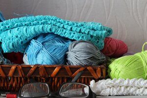 Ein geflochtener Handarbeitskorb gef&uuml;llt mit verschiedenen bunten Wollkn&auml;ueln und einem geh&auml;kelten, t&uuml;rkisfarbenen Tuch, davor liegen eine Lesebrille und ein wei&szlig;es Strickst&uuml;ck.