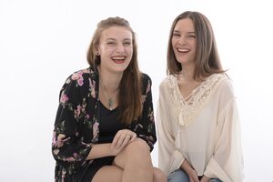 Zwei junge Frauen sitzen nebeneinander vor einem schlichten, rein wei&szlig;en Hintergrund und lachen herzlich in die Kamera. Die linke Frau hat hellbraunes, teilweise hochgestecktes Haar und tr&auml;gt einen schwarzen Kimono mit einem bunten Blumenmuster &uuml;ber einem dunklen Top sowie zwei Halsketten. Die rechte Frau hat lange, glatte braune Haare und tr&auml;gt eine lockere, cremefarbene Bluse mit Spitzenbesatz am Ausschnitt und Kordeln. Beide strahlen eine ausgelassene, fr&ouml;hliche Stimmung aus.