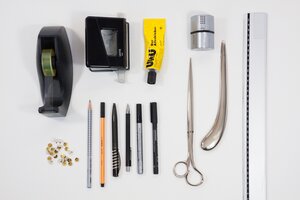 Eine Draufsicht (Flatlay) auf verschiedene B&uuml;ro- und Bastelutensilien, die ordentlich auf einem rein wei&szlig;en Untergrund nebeneinander aufgereiht sind.

In der oberen Reihe befinden sich von links nach rechts ein schwarzer Klebebandabroller, ein schwarzer Leitz-Locher, eine gelbe Tube UHU-Alleskleber und ein kleiner zylindrischer, silberner Anspitzer.

Darunter liegt eine Reihe von Stiften und Kleinteilen: Ganz links ein kleiner Haufen Rei&szlig;zwecken, gefolgt von einem grau gepunkteten Bleistift, einem orangefarbenen Stabilo-Fineliner, zwei schwarzen Kugelschreibern und zwei schwarzen Filzstiften (darunter ein Staedtler Lumocolor).

Rechts daneben sind eine lange, silberne Metallschere sowie ein geschwungenes, silbernes Falzbein platziert. Den rechten Bildrand schlie&szlig;t ein langes, transparentes Kunststofflineal vertikal ab.