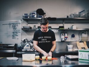 Ein junger Mann mit kurzen Haaren und Bart steht in einer professionellen K&uuml;che an einer Edelstahl-Arbeitsfl&auml;che und bereitet Speisen zu. Er blickt konzentriert nach unten und bef&uuml;llt mehrere durchsichtige Plastikbecher, die nebeneinander auf einem roten Schneidebrett stehen. Er tr&auml;gt ein schwarzes T-Shirt mit der wei&szlig;en Aufschrift &raquo;COPS & ROBBERS SMASH COWBOYS & INDIANS&laquo;. Im Hintergrund sind K&uuml;chenger&auml;te wie eine Aufschnittmaschine, Edelstahlregale mit Boxen und ein Whiteboard mit Notizen zu sehen.