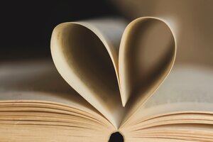 Eine Nahaufnahme eines aufgeschlagenen Buches mit vergilbten Seiten. Zwei gegen&uuml;berliegende Buchseiten sind zur Mitte hin so eingeknickt und zusammengesteckt, dass sie von vorne betrachtet die Form eines dreidimensionalen Herzens bilden. Der Fokus liegt scharf auf der Herzform im Vordergrund, w&auml;hrend die restlichen Buchseiten und der dunkle Hintergrund in eine weiche Unsch&auml;rfe verlaufen. Die Beleuchtung ist warm und stimmungsvoll.
