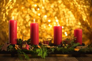 Ein l&auml;ngliches Adventsgesteck auf einer schwarzen Schale, platziert auf einem Holztisch. Von den vier gro&szlig;en, roten Stumpenkerzen brennen die linken drei mit einer warmen Flamme, w&auml;hrend die ganz rechte Kerze unangez&uuml;ndet ist (Symbol f&uuml;r den 3. Advent). Das Gesteck ist festlich mit gr&uuml;nen Tannenzweigen, Thuja, Efeu, Zimtstangen, getrockneten Orangenscheiben, kleinen roten Geschenkp&auml;ckchen und goldenen Perlen dekoriert. Der Hintergrund besteht aus einem stark verschwommenen, golden glitzernden Lichtermeer mit warmem Bokeh-Effekt.