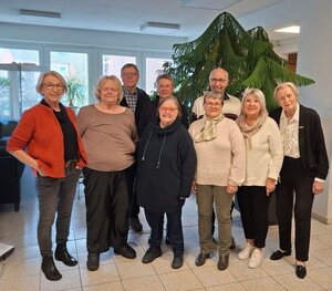 Bild vergr&ouml;&szlig;ern: Das Foto zeigt eine Gruppe von neun &auml;lteren Erwachsenen (sieben Frauen und zwei M&auml;nner), vermutlich der Seniorenbeirat der Stadt Geesthacht. Sie stehen in einem hellen, gefliesten Innenraum eng nebeneinander und l&auml;cheln in die Kamera. Im Hintergrund befindet sich eine gro&szlig;e Fensterfront, die den Blick nach drau&szlig;en andeutet, sowie eine sehr gro&szlig;e, &uuml;ppige gr&uuml;ne Zimmerpflanze (Zimmertanne), die hinter der Gruppe aufragt.