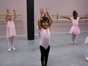 Eine Gruppe kleiner M&auml;dchen &uuml;bt in einem Ballettsaal mit grauem Boden und zartrosa W&auml;nden. Im Hintergrund sind Ballettstangen an der Wand montiert.

Im Vordergrund steht ein junges M&auml;dchen mit dunklen Haaren und einer rosa Haarschleife, das konzentriert beide Arme nach oben streckt. Sie tr&auml;gt ein rosa Balletttrikot und eine lange, schwarze Leggings. Links im Hintergrund steht ein weiteres M&auml;dchen in einem rosa Ballettkleidchen mit erhobenen Armen. Rechts im Hintergrund streckt ein dunkelh&auml;utiges M&auml;dchen im klassischen rosa Tutu und wei&szlig;en Strumpfhosen die Arme seitlich aus. In der Mitte des Raumes steht eine dicke, schwarze S&auml;ule.