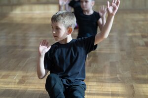 Innenaufnahme aus einer leicht erh&ouml;hten Perspektive, die Kinder beim Tanz- oder Gymnastiktraining in einem Tanzsaal zeigt. Im scharfen Vordergrund sitzt ein kleiner Junge mit kurzen, blonden Haaren in schwarzer Sportkleidung auf dem hellen Holzparkettboden. Er hat den linken Arm nach oben angewinkelt, die Handfl&auml;che zeigt nach vorne, w&auml;hrend er konzentriert nach links blickt. Hinter ihm sitzt versetzt ein M&auml;dchen, ebenfalls in schwarzer Kleidung, und f&uuml;hrt dieselbe Bewegung synchron aus. Der Hintergrund des Saals verl&auml;uft in einer weichen Unsch&auml;rfe, wodurch das rhythmische Training im Fokus steht.