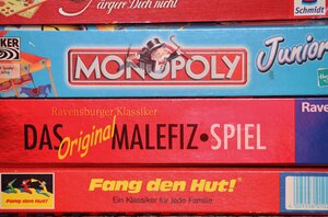 Eine bunte Nahaufnahme aus der Vogelperspektive zeigt eine Sammlung verschiedener, &uuml;bereinandergestapelter Gesellschaftsspiele und Schachteln. Ganz oben liegt ein kleineres, gelb-orangefarbenes Kartenspiel mit der gro&szlig;en Aufschrift &raquo;6 nimmt!&laquo;. Darunter sind Teile von gr&ouml;&szlig;eren Spieleboxen zu sehen, darunter das taktische Legespiel &raquo;Carcassonne&laquo; (erkennbar an der typischen mittelalterlichen Burg-Illustration) sowie ein weiteres Spiel mit einer Grafik, die eine historische europ&auml;ische Stadt oder Festung an einer K&uuml;ste zeigt. Die Schachteln liegen leicht versetzt, sodass die unterschiedlichen farbigen Cover und Designs eine dynamische, spielerische Collage bilden.