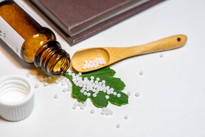 Eine Nahaufnahme aus der Vogelperspektive auf ein hom&ouml;opathisches Arrangement auf wei&szlig;em Untergrund. Ein kleines, braunes Medizinfl&auml;schchen aus Glas liegt umgekippt auf der linken Seite, wobei zahlreiche wei&szlig;e Globuli-Streuk&uuml;gelchen aus der &Ouml;ffnung herausgerollt sind. Viele der K&uuml;gelchen liegen auf einem einzelnen, gr&uuml;nen, gezackten Blatt im Zentrum des Bildes. Daneben befindet sich ein kleiner, schmaler Holzl&ouml;ffel, der ebenfalls mit einigen Globuli gef&uuml;llt ist. Im linken Vordergrund ist der abgeschraubte, wei&szlig;e Kunststoffdeckel des Fl&auml;schchens unscharf zu sehen, w&auml;hrend im oberen Hintergrund die Ecke eines geschlossenen, braunen Buches ins Bild ragt.