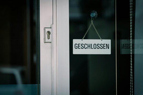 Nahaufnahme einer gl&auml;sernen Eingangst&uuml;r mit einem wei&szlig;en Hinweisschild. Das Schild h&auml;ngt an einer feinen Metallkette, die mit einem Saugnapf an der dunklen Glasscheibe befestigt ist, und tr&auml;gt die Aufschrift &raquo;GESCHLOSSEN&laquo; in schwarzen Gro&szlig;buchstaben. Links im Bild ist der wei&szlig;e T&uuml;rrahmen mit einem Schl&uuml;sselloch zu sehen. Auf der rechten Seite spiegelt sich der Text des Schildes spiegelverkehrt in einer parallel verlaufenden Glasscheibe, neben der eine wei&szlig;e Kugelkette eines Rollos herunterh&auml;ngt.