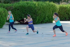 Drei Frauen machen gemeinsam Sport im Freien auf einem gepflasterten Platz vor einer gro&szlig;en, dichten gr&uuml;nen Hecke. Sie befinden sich alle in der Ausfallschritt-Position (Lunge). Die linke und die rechte Frau tragen jeweils ein t&uuml;rkisfarbenes Top und eine dunkle Leggings, w&auml;hrend die Frau in der Mitte ein lila Sport-Bustier und gemusterte, kurze Leggings tr&auml;gt. Auf dem Boden im Vordergrund steht eine kleine Wasserflasche.