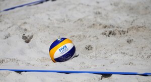 Ein offizieller Beachvolleyball der Marke Mikasa liegt im feink&ouml;rnigen, hellen Sand eines Beachvolleyballfeldes. Der Ball hat das charakteristische geschwungene Design in den Farben Blau, Gelb und Wei&szlig;. Auf dem wei&szlig;en Segment prangt das blaue Logo des internationalen Volleyballverbandes &raquo;FIVB&laquo; mit dem Zusatz &raquo;OFFICIAL GAME BALL&laquo;. Auf einem blauen Segment dar&uuml;ber ist zudem das gelb-blaue Logo des europ&auml;ischen Verbandes &raquo;CEV&laquo; aufgedruckt. Der Ball liegt leicht versenkt im Sand, der im Vordergrund durch eine blaue, flach gespannte Begrenzungslinie markiert wird. Der Hintergrund zeigt die leicht unebene Sandstruktur und ist dezent unscharf gehalten.