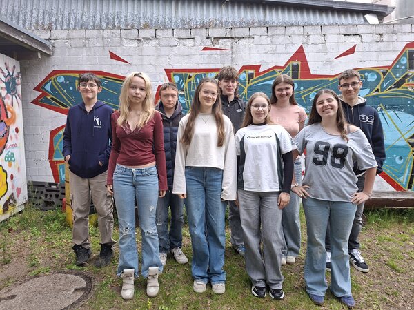 Das Foto zeigt eine Gruppe von acht Jugendlichen - vier Jungen und vier M&auml;dchen - im Alter von etwa 13 bis 17 Jahren. Sie stehen in zwei Reihen nebeneinander im Freien vor einer wei&szlig; gestrichenen Betonmauer, die gro&szlig;fl&auml;chig mit einem bunten Graffiti in den Farben Blau, Rot, Gelb und T&uuml;rkis bespr&uuml;ht ist. Der Boden im Vordergrund besteht aus unebenem Gras und etwas Erde; unten links ist ein runder Gullydeckel aus Beton zu sehen. Die Jugendlichen blicken alle freundlich oder l&auml;chelnd direkt in die Kamera.