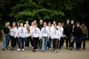 Eine gro&szlig;e Gruppe von Menschen unterschiedlichen Alters l&auml;uft gemeinsam entschlossen nach vorne auf die Kamera zu. Der Gro&szlig;teil der Gruppe tr&auml;gt einheitliche wei&szlig;e Kapuzenpullover, auf denen auf der Brust ein gro&szlig;es, geschwungenes, schwarzes &raquo;R&laquo; gedruckt ist. Einige Personen im Hintergrund und am rechten Rand tragen dunkle Kleidung; ganz rechts h&auml;lt jemand Musikequipment. Im Hintergrund befindet sich eine dichte, gr&uuml;ne Wand aus B&auml;umen und Geb&uuml;sch.