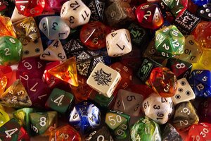 Eine bunte und dichte Nahaufnahme einer gro&szlig;en Sammlung verschiedenster Spielw&uuml;rfel (haupts&auml;chlich polyedrische Rollenspielw&uuml;rfel wie W4, W6, W10, W12 und W20). Die W&uuml;rfel haben unterschiedlichste Farben, Muster und Effekte - darunter marmorierte, transparente, glitzernde und einfarbige Designs in Rot, Gr&uuml;n, Blau, Gold, Wei&szlig; und Schwarz. Zentral im Fokus liegt ein cremefarbener, sechsseitiger W&uuml;rfel, auf dessen Oberseite statt einer Zahl ein schwarzes Symbol eines z&auml;hnefletschenden Wolfskopfes (das Wolfs-Medaillon aus The Witcher) abgebildet ist. Die Zahlen auf den umliegenden W&uuml;rfeln sind in verschiedenen Schriftarten und Farben wie Wei&szlig;, Gold oder Silber eingepr&auml;gt.