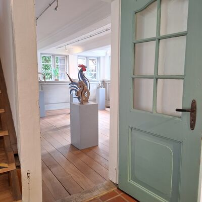 Blick durch eine ge&ouml;ffnete, altmodische Holzt&uuml;r in Salbeigr&uuml;n mit Glaseins&auml;tzen in einen hellen Ausstellungsraum. Im Vordergrund sind braune Bodenfliesen und links ein Ansatz einer h&ouml;lzernen Treppe zu sehen. Der dahinterliegende Raum verf&uuml;gt &uuml;ber einen warmen Holzdielenboden, wei&szlig;e W&auml;nde und eine niedrige Decke mit Lichtschienen. Zentral im Raum steht eine moderne, skulpturale Kunstnahe eines Hahns auf einem grauen, quaderf&ouml;rmigen Sockel. Im Hintergrund sind weitere Sockel mit Ausstellungsst&uuml;cken vor Sprossenfenstern platziert, die den Blick ins Gr&uuml;ne freigeben.