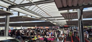Belebter Flohmarkt unter den halbtransparenten &Uuml;berdachungen eines Parkplatzes vor Gesch&auml;ften. Zahlreiche Besucher st&ouml;bern an Tischen mit Haushaltswaren und St&auml;ndern voll Kleidung.