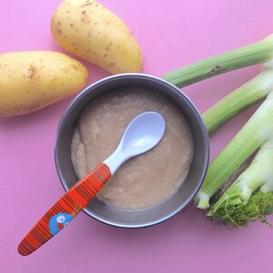 Eine graue Sch&uuml;ssel mit Babybrei steht auf rosa Hintergrund. In der Sch&uuml;ssel liegt ein roter Babyl&ouml;ffel. Neben der Sch&uuml;ssel befinden sich zwei gelbe Kartoffeln und frischer Fenchel.