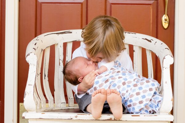 Bild vergr&ouml;&szlig;ern: Ein Kleinkind sitzt auf einer wei&szlig;en Holzbank und h&auml;lt ein schlafendes Baby im Arm. Es gibt dem Baby einen z&auml;rtlichen Kuss auf das Gesicht.