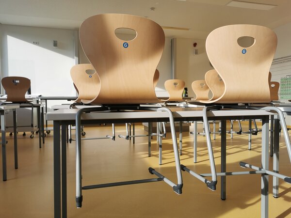 In einem hellen Klassenzimmer stehen St&uuml;hle aus Holz umgedreht auf den Tischen. Die St&uuml;hle haben Beine aus grauem Metall und sind mit einer blauen Zahl sechs markiert. Warmes Sonnenlicht scheint in den Raum.