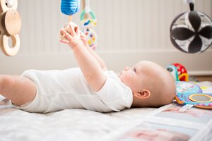 Ein Baby liegt auf dem R&uuml;cken und greift aufmerksam mit beiden H&auml;nden nach einem h&auml;ngenden Spielzeug. Es tr&auml;gt einen wei&szlig;en Body.