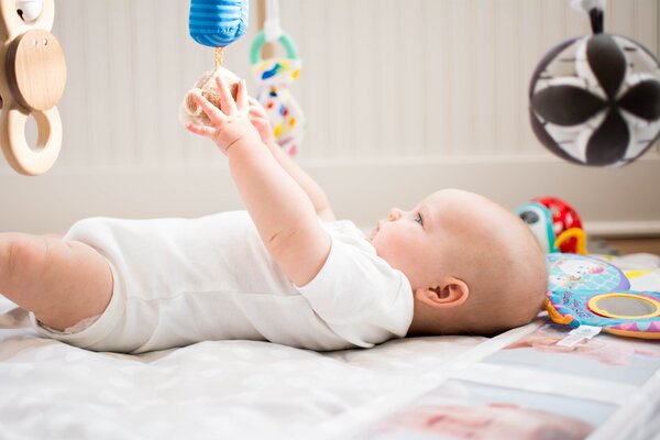 Bild vergr&ouml;&szlig;ern: Ein Baby liegt auf dem R&uuml;cken und greift aufmerksam mit beiden H&auml;nden nach einem h&auml;ngenden Spielzeug. Es tr&auml;gt einen wei&szlig;en Body.