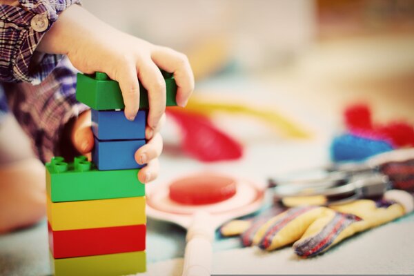 Eine Kinderhand stapelt bunte Bausteine zu einem Turm. Die Steine sind rot, gelb, gr&uuml;n und blau. Im Hintergrund liegen weitere Spielsachen.