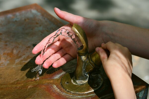 Zwei H&auml;nde bedienen einen Trinkbrunnen im Freien. Aus dem Messinghahn flie&szlig;t ein hoher Wasserstrahl. Eine Hand f&auml;ngt das frische Wasser auf.