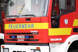 Nahaufnahme von der roten Front eines Feuerwehrautos der Marke Iveco Magirus. Auf einem gelben Streifen steht in blauen Buchstaben das Wort Feuerwehr. Das Fahrzeug besitzt blaue Blitzer am K&uuml;hlergrill und eine gro&szlig;e Windschutzscheibe.
