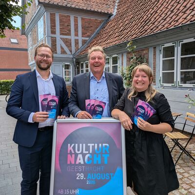 Drei l&auml;chelnde Personen stehen vor einem historischen Fachwerkhaus und halten Flyer f&uuml;r die Kulturnacht Geesthacht in der Hand. Vor ihnen steht ein gro&szlig;es Plakat zur Kulturnacht Geesthacht am 29. August.