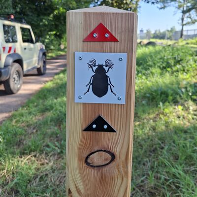Ein Holzpfosten steht an einem Weg. Auf ihm ist ein Schild mit einem schwarzen K&auml;fer. Dar&uuml;ber ist ein rotes Dreieck angebracht, darunter ein schwarzes. Im Hintergrund steht ein Gel&auml;ndewagen auf einem Weg.