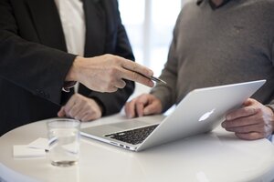 Zwei M&auml;nner sitzen zusammen an einem wei&szlig;en Tisch. Einer zeigt mit einem Stift auf einen ge&ouml;ffneten Laptop. Vor ihnen steht ein Glas Wasser.