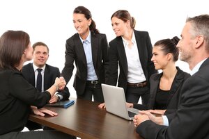 Eine Gruppe von Gesch&auml;ftsleuten in dunkler Kleidung sitzt und steht an einem Konferenztisch. Eine stehende Frau gibt einer Person am Tisch die Hand. Auf dem Tisch steht ein wei&szlig;er Laptop. Alle schauen freundlich.