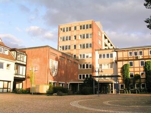 Ein gro&szlig;es Geb&auml;ude aus rotem Backstein mit einem Wappen an der Fassade. Davor befindet sich ein gepflasterter Platz mit Laternen unter einem leicht bew&ouml;lkten Himmel.