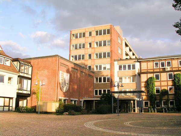 Ein gro&szlig;es Geb&auml;ude aus rotem Backstein mit einem Wappen an der Fassade. Davor befindet sich ein gepflasterter Platz mit Laternen unter einem leicht bew&ouml;lkten Himmel.