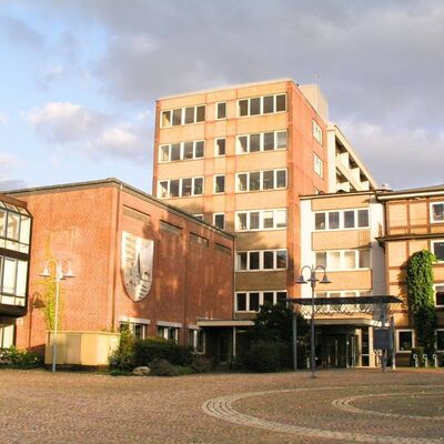 Ein gro&szlig;es Geb&auml;ude aus rotem Backstein mit einem Wappen an der Fassade. Davor befindet sich ein gepflasterter Platz mit Laternen unter einem leicht bew&ouml;lkten Himmel.