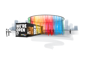 Ein schwarzer Ausstellungs-Container steht im Freien. Darauf steht in wei&szlig;er Schrift WE'RE OPEN!? und WIE TOLERANT BIST DU?. Daneben h&auml;ngt ein gro&szlig;er, runder Vorhang aus vielen bunten B&auml;ndern in Regenbogen-Farben.