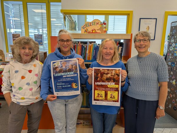 Vier l&auml;chelnde Frauen stehen in einer Bibliothek vor einem bunten B&uuml;cherregal. Die beiden Frauen in der Mitte pr&auml;sentieren Plakate f&uuml;r den Vorleseclub Lesefreu(n)de.