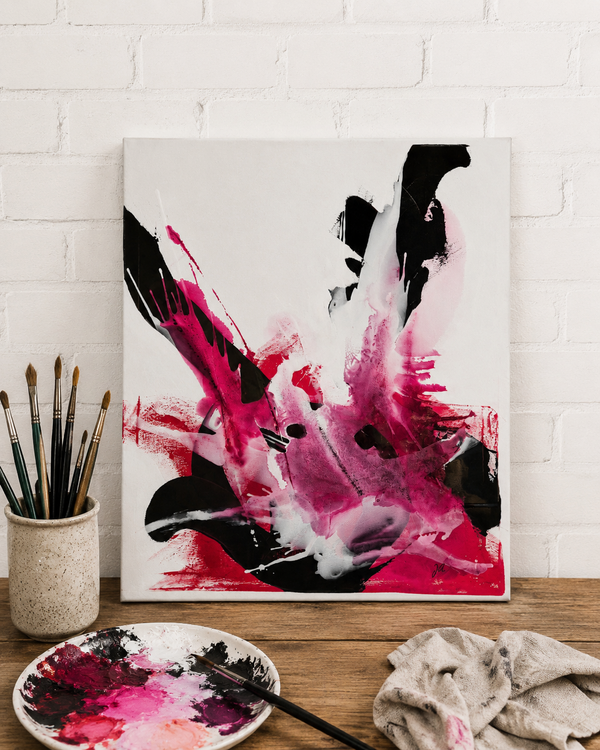 Bild vergr&ouml;&szlig;ern: Ein abstraktes Bild lehnt an einer wei&szlig;en Ziegelwand auf einem Holztisch. Es zeigt dynamische Farbspritzer in Pink, Magenta und Schwarz. Links steht ein Becher voller Pinsel.