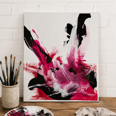 Ein abstraktes Bild lehnt an einer wei&szlig;en Ziegelwand auf einem Holztisch. Es zeigt dynamische Farbspritzer in Pink, Magenta und Schwarz. Links steht ein Becher voller Pinsel.