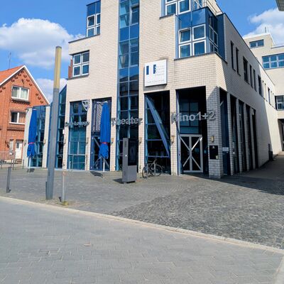 Ein modernes, helles Geb&auml;ude mit vielen blauen Glasfenstern bei Sonnenschein. Auf der Fassade steht Theater und Kino 1+2. Vor dem Eingang stehen blaue Sonnenschirme auf einem gepflasterten Platz.